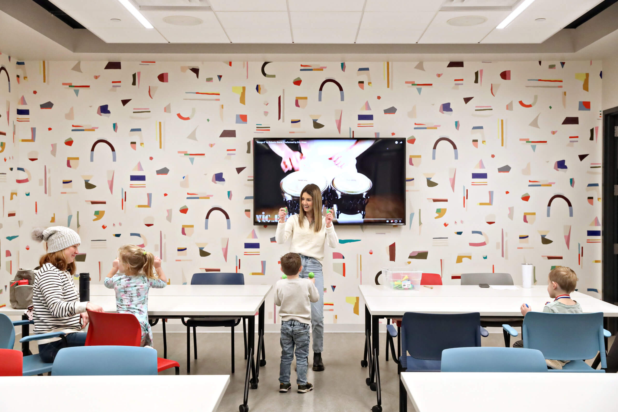 An instructor leading a class for young children in a colorful classroom with abstract wall designs.