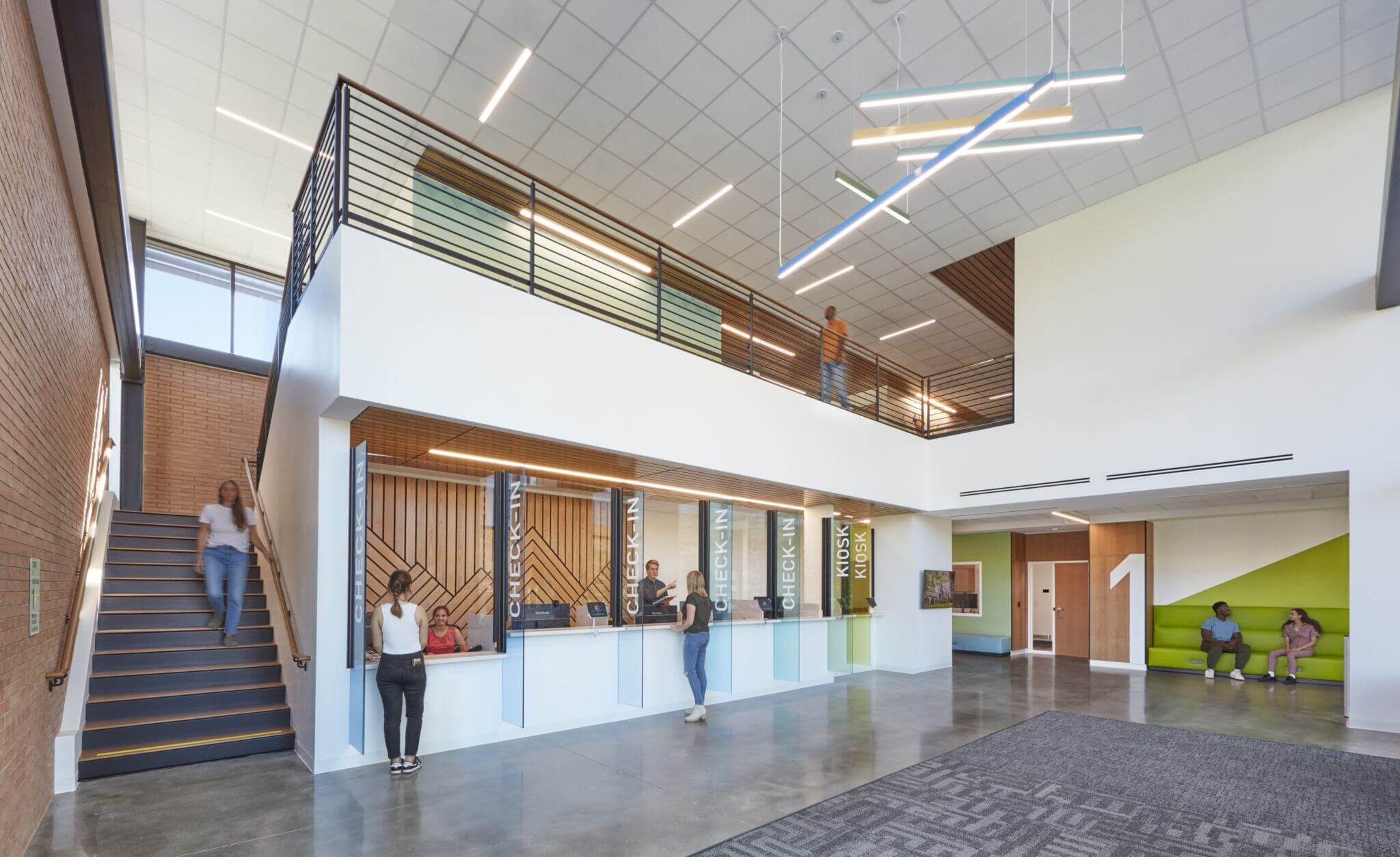 Modern school lobby with people ascending and descending a staircase, a reception desk with two individuals, and others sitting and conversing in a lounging area. The interior features large windows, high ceilings with distinctive hanging lights, and a combination of wooden and metal accents.