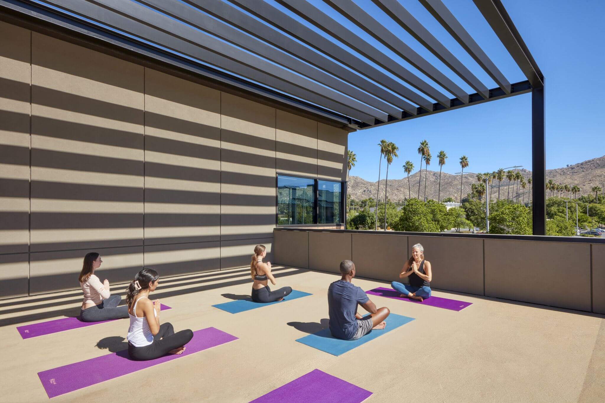 A yoga class takes place outdoors under a large modern pergola, with participants seated on colorful mats, facing an instructor with a scenic backdrop of palm trees and mountains.