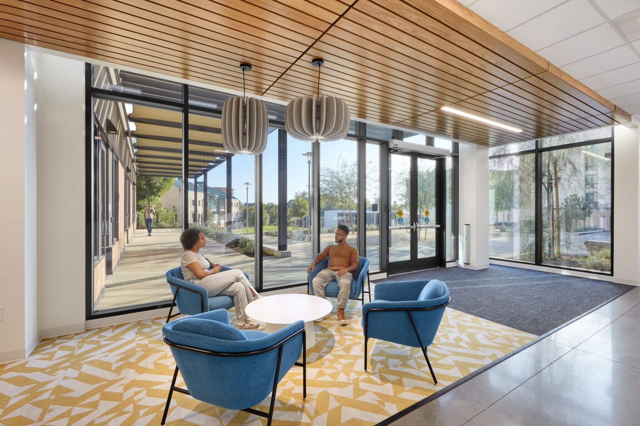 Modern office lobby with floor-to-ceiling windows showing two people seated in blue armchairs engaged in conversation. The interior features a wooden slatted ceiling, geometric patterned carpet, and futuristic hanging light fixtures.