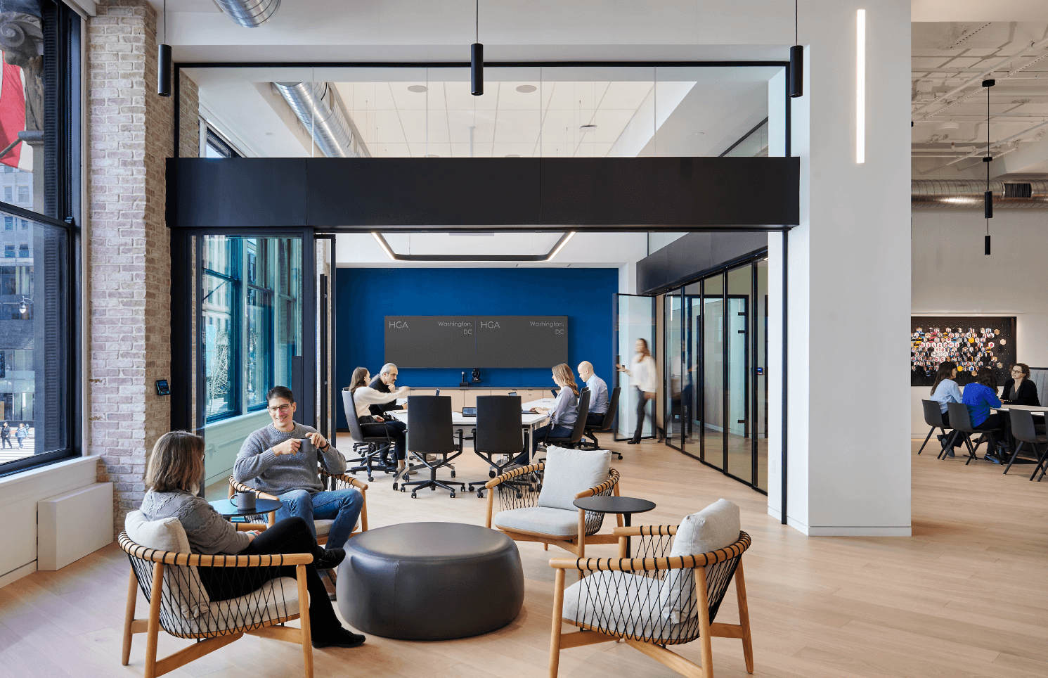 Interior view of a spacious office with people working at desks and conversing in a lounge area. The room features modern decor with large windows, exposed brick walls, and a prominent blue accent wall with a flat-screen TV.