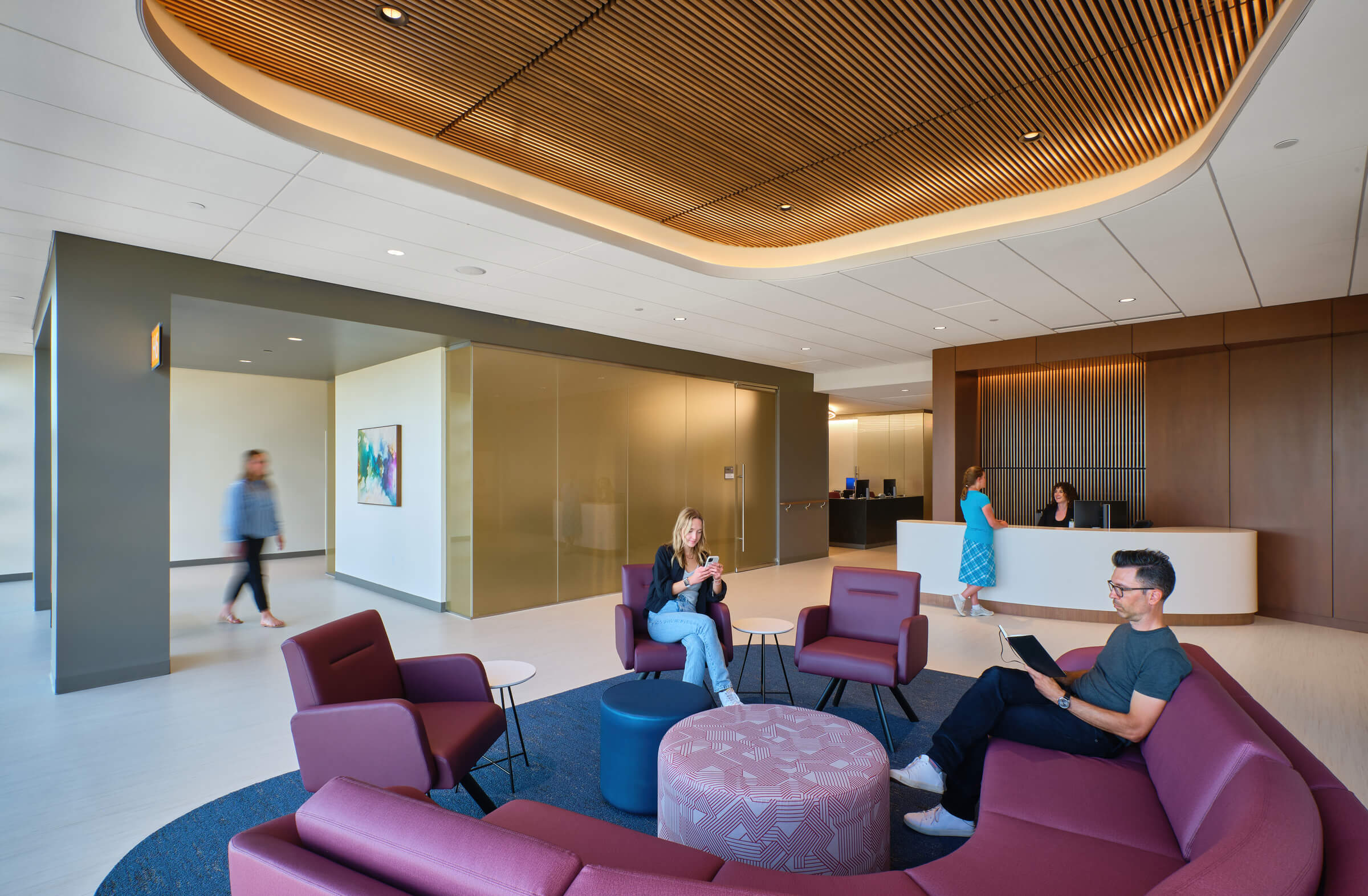 Interior of a modern office lobby featuring a reception desk, several individuals seated on sofas and chairs, and others walking. The design includes warm wood tones and a curved ceiling.