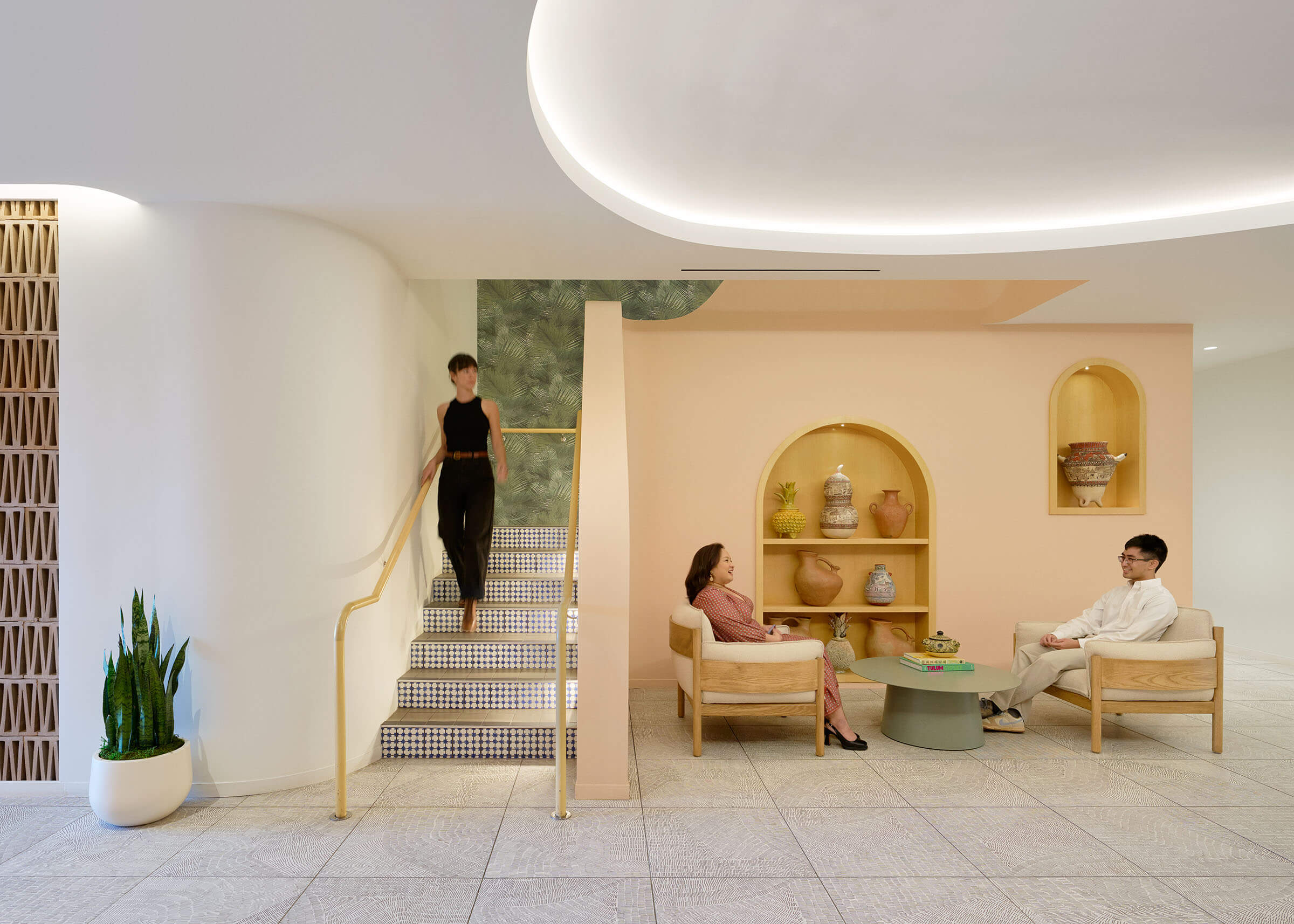 Interior of a modern office lounge with three individuals engaged in conversation and activities. The room features soft peach walls, stylish curved lighting overhead, and minimalist furniture. One person descends a stairway while two others sit across from each other, conversing beside a table with books and decorative items.