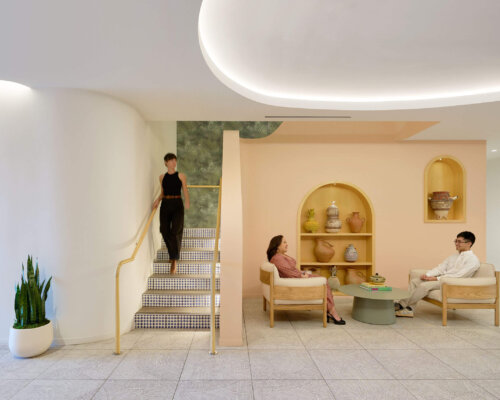 Interior of a modern office lounge with three individuals engaged in conversation and activities. The room features soft peach walls, stylish curved lighting overhead, and minimalist furniture. One person descends a stairway while two others sit across from each other, conversing beside a table with books and decorative items.