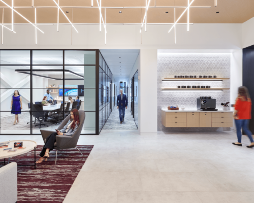 Interior of a modern office space featuring a sleek reception desk, overhead geometric lighting fixtures, and people engaged in various activities. One individual is seated near the entrance, another is walking towards a hallway, and a third is interacting near a work area with digital screens.