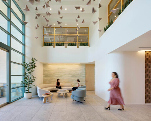 Interior of a modern office lobby with three individuals seated and conversing. The décor features geometric hanging elements, a two-level layout with balconies, and minimalistic furniture. A person in motion blurs in the foreground.