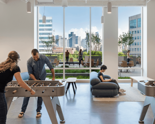 Two individuals playing foosball in a modern office space with large windows showcasing a cityscape. Another person is seated in the background using a laptop.