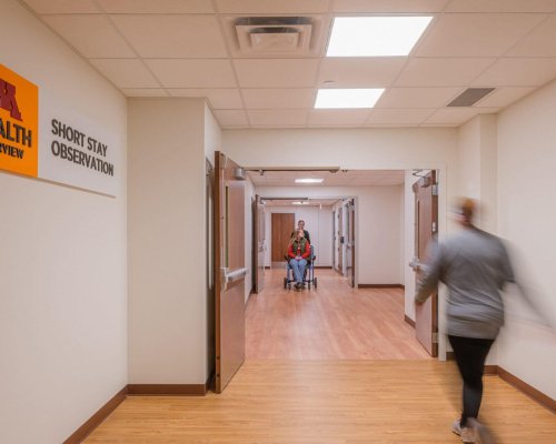 Hospital hallway where a patient walks past a sign reading M Health Fairview Short Stay Observation