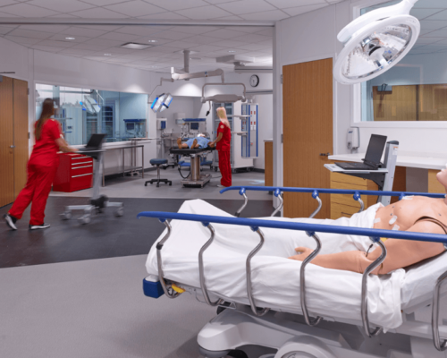 title image of nursing lab with a dummy patient and nurses in red scrubs
